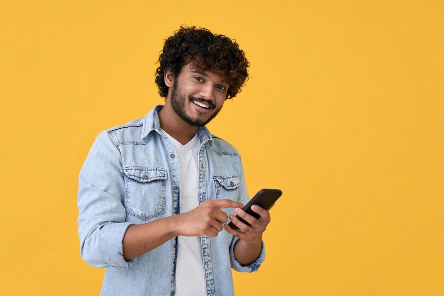 Young man in denim jacket smiling while searching for rentals on his phone